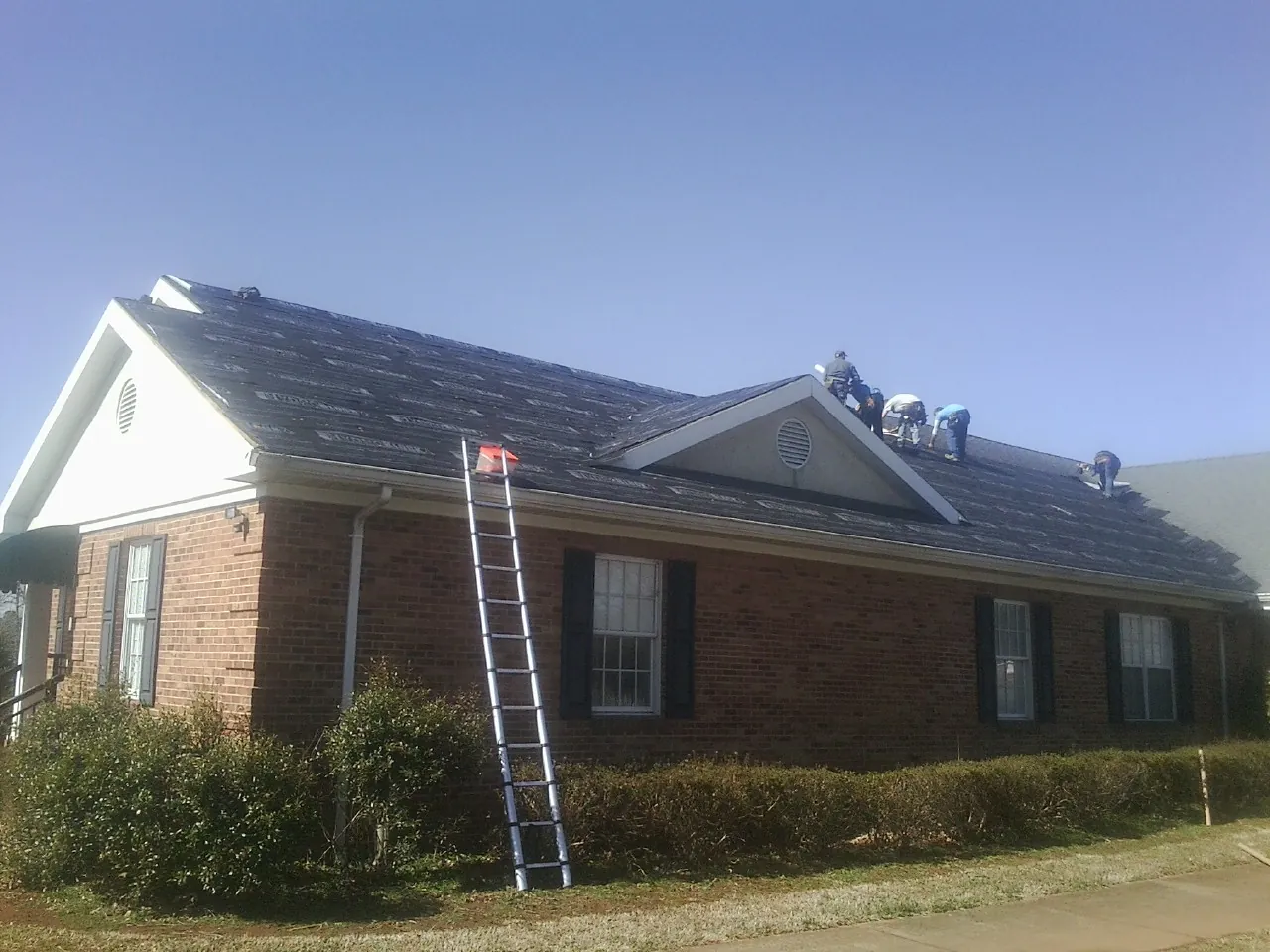 Crew tearing shingles off a brick ranch roof with a ladder set — Emergency Roof Repair work by our crew in Gasburg