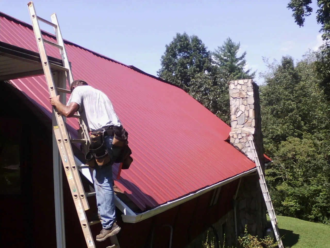 Roofer on a ladder beside a red metal roof and stone chimney — Soffit & Fascia Repair by Roofing Brothers in Gasburg